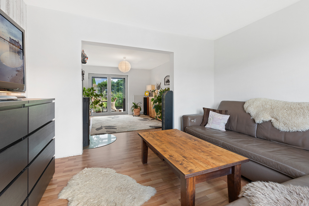 Wohnzimmer mit braunem Ledersofa, Holztisch und Blick auf einen hellen Raum mit Terrassentür.