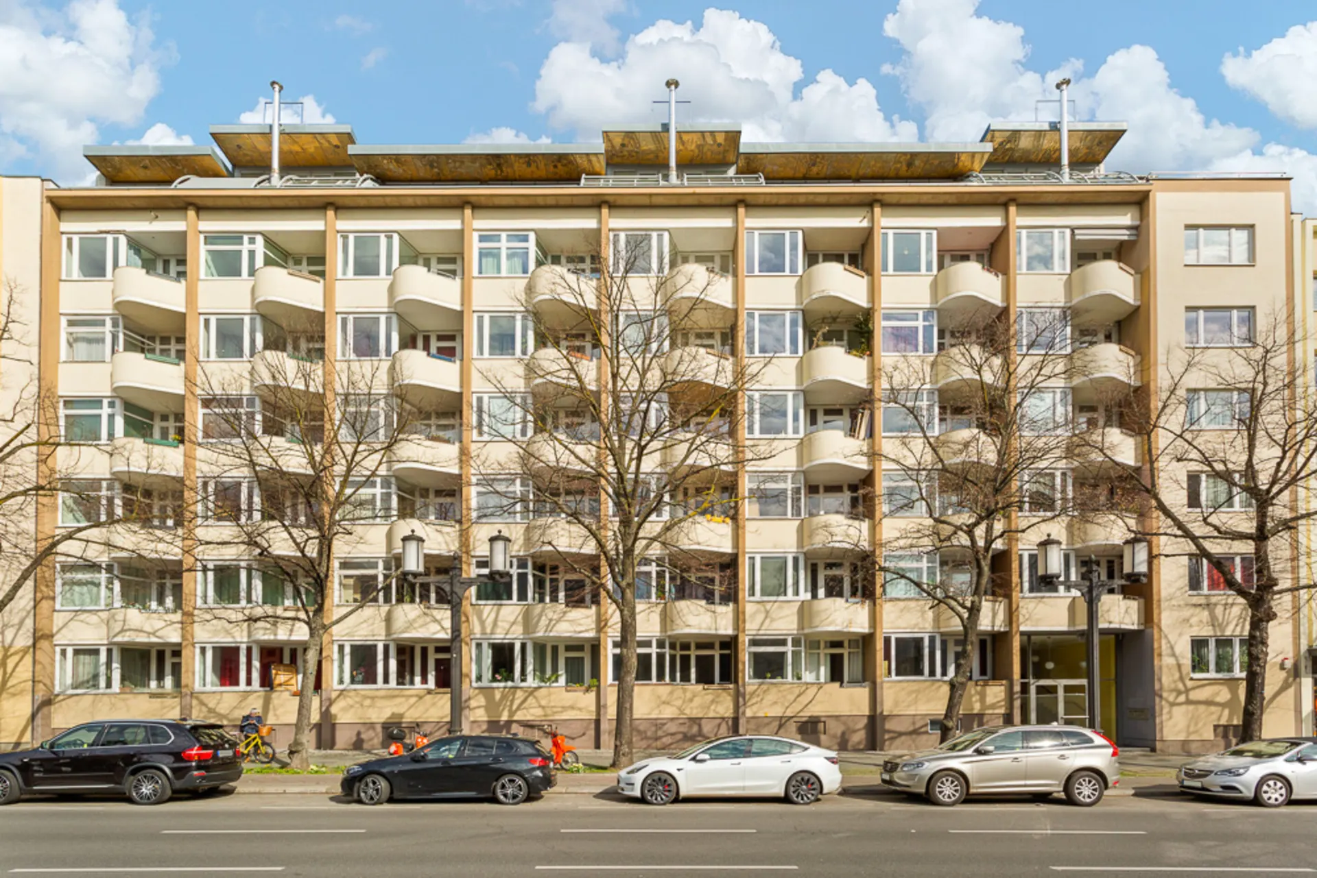 Mehrstöckiges Wohnhaus mit Balkonen, geparkten Autos und Bäumen davor.