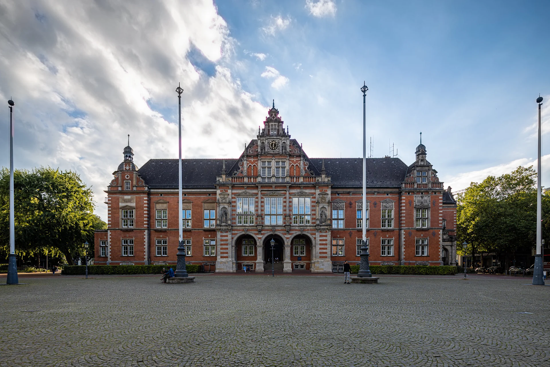 Großes, historisches Backsteingebäude mit Uhr und Verzierungen, davor gepflasterter Platz.