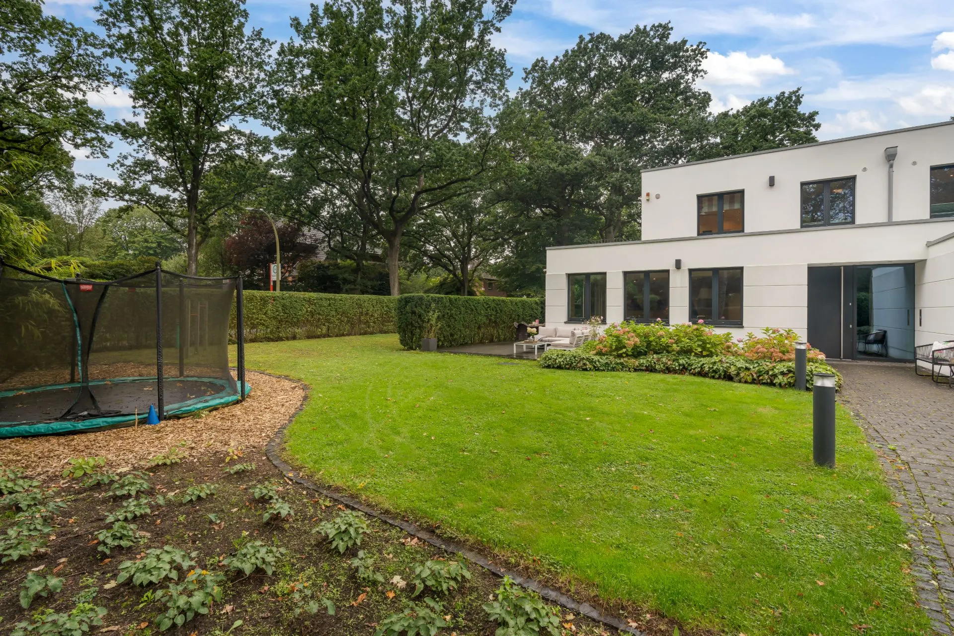 Moderner Garten mit Rasen, Trampolin, Terrasse und weißem Wohnhaus im Hintergrund.