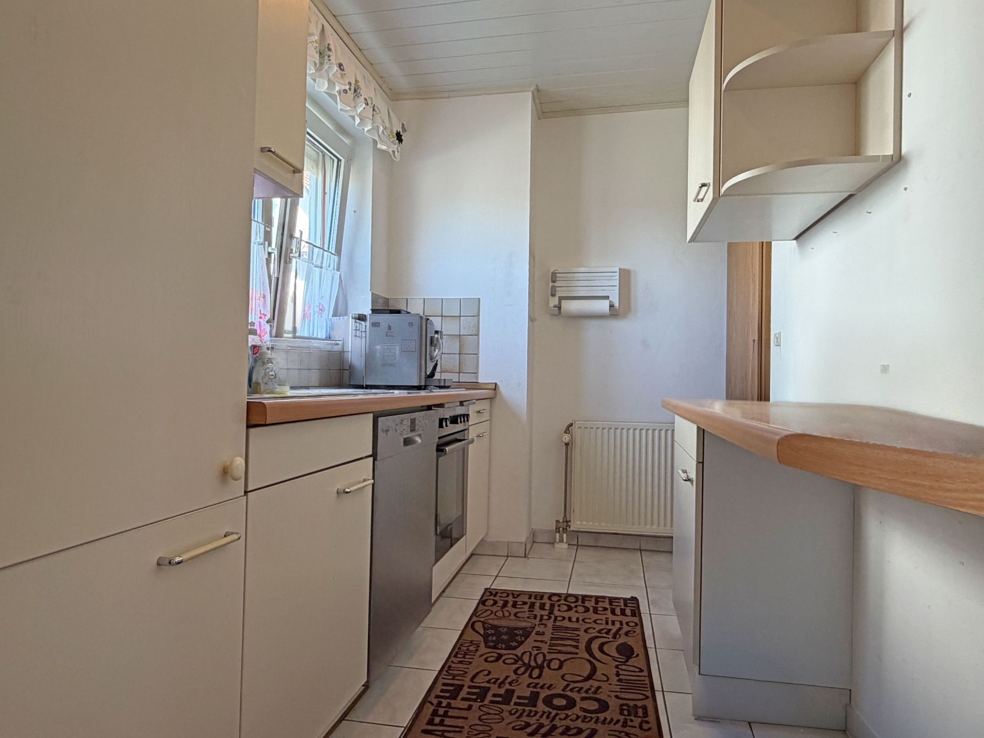 Helle Küche mit weißen Schränken, Arbeitsplatte, Fenster, Spülmaschine und Regalwand.
