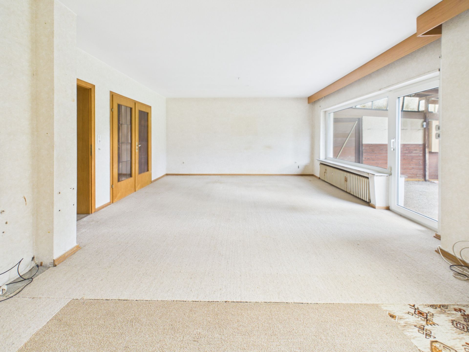 Leeres Wohnzimmer mit Teppichboden, Holztüren und großem Fenster zur Terrasse.