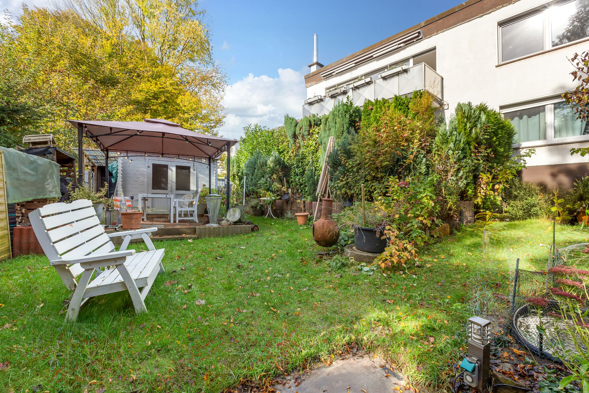 Grüner Garten mit Sitzbank, Pavillon und vielen Pflanzen neben einem Wohnhaus.