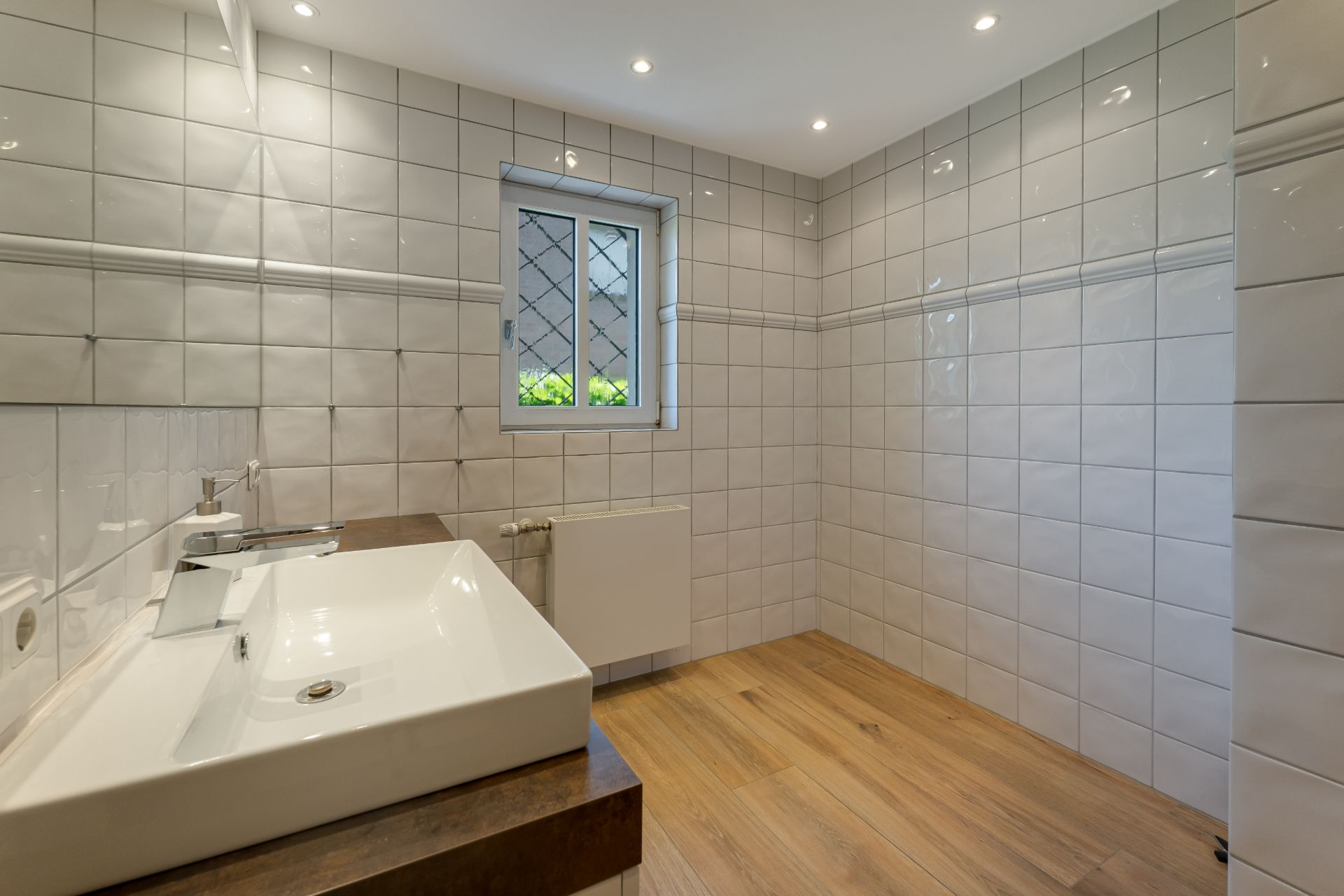 Modernes Bad mit weiß gefliesten Wänden, Holzboden, Waschbecken und Fenster mit Gitter.