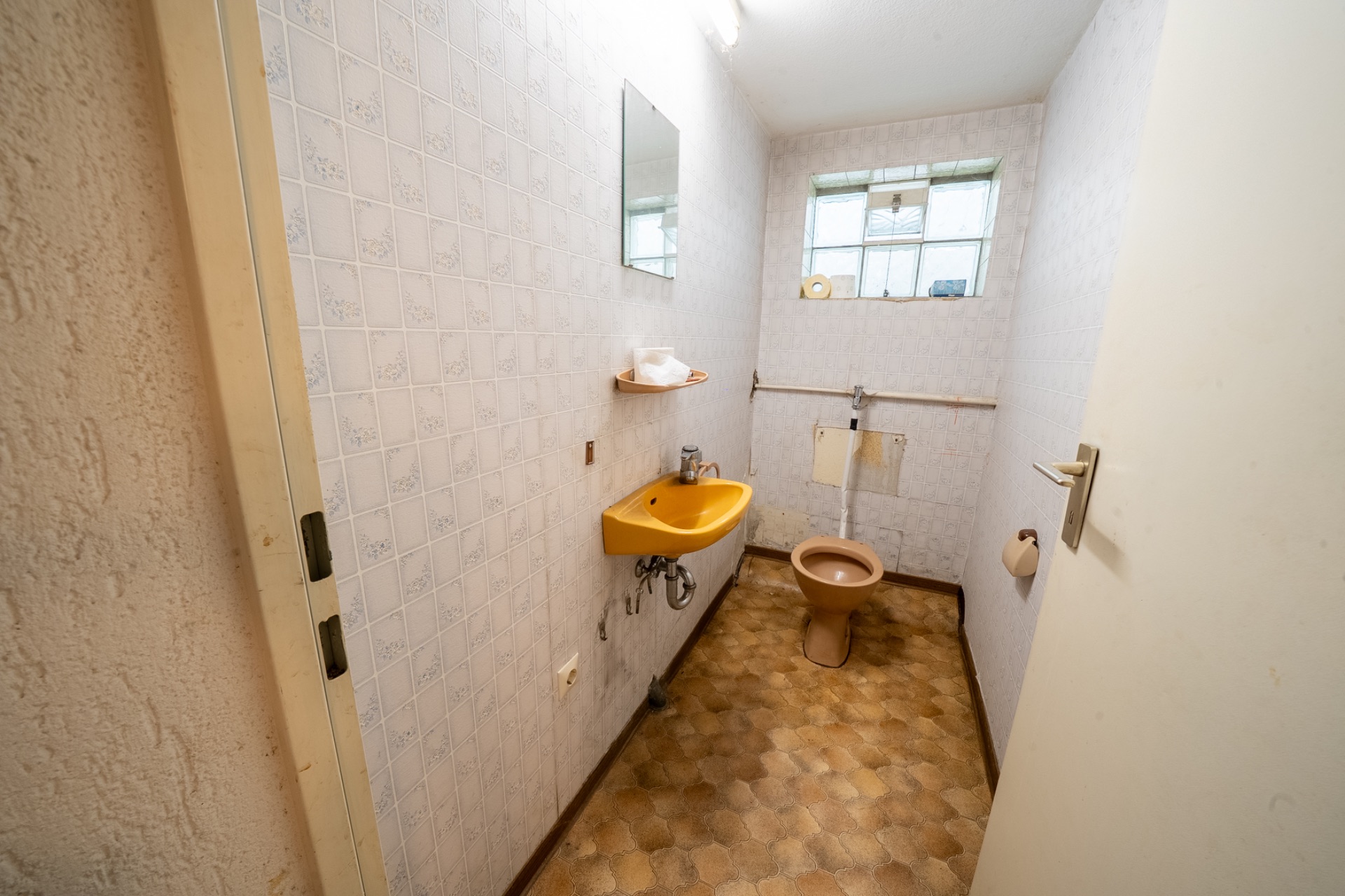 Schmales Bad mit gelbem Waschbecken, brauner Toilette, Spiegel und Fenster.
