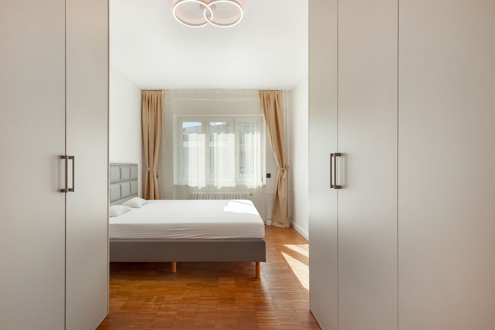 Helles Schlafzimmer mit Doppelbett, Holzboden, Kleiderschränken und Fenster mit Vorhängen.