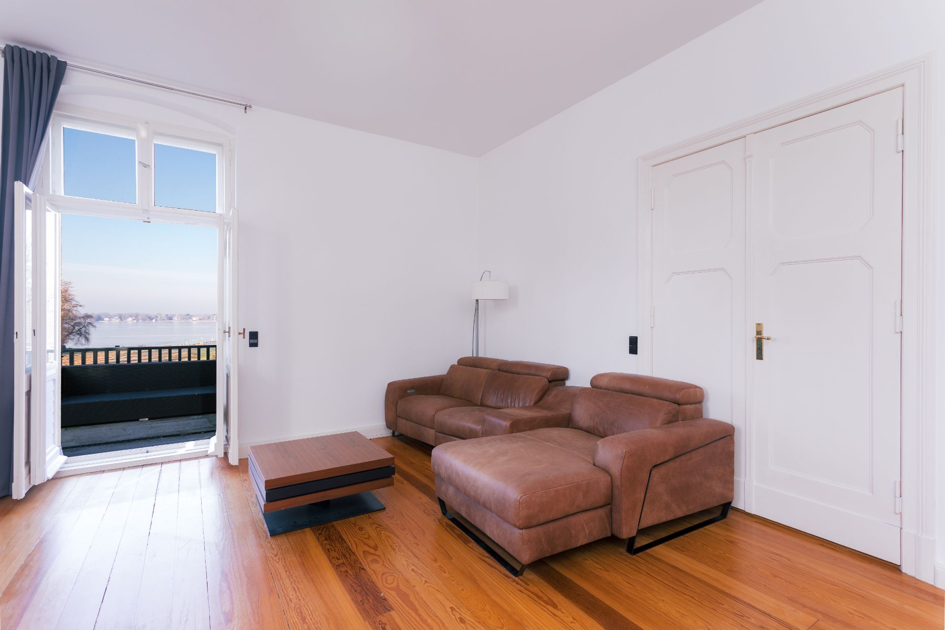Wohnzimmer mit braunem Sofa, Holztisch, Parkettboden und offenem Balkon mit Ausblick.
