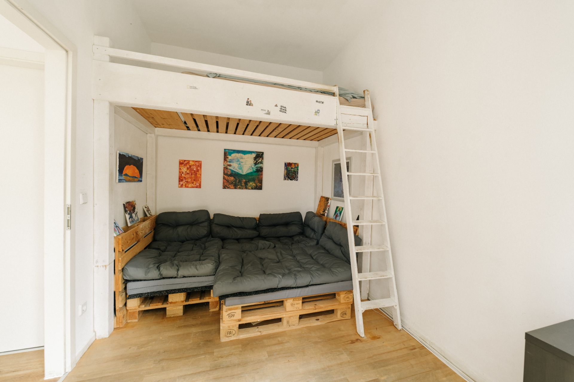 Wohnzimmer mit Palettensofa, Hochbett und Leiter, helle Wände, Holzboden, Wandbilder.