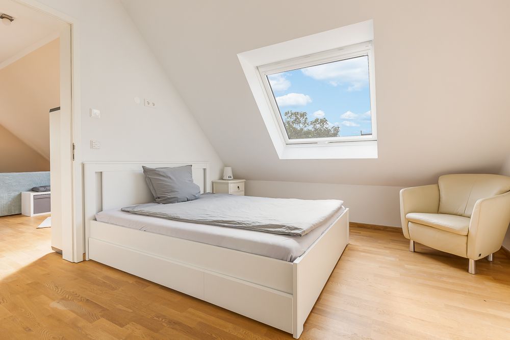 Helles Schlafzimmer mit Einzelbett, grauem Kissen, Sessel und Dachfenster.
