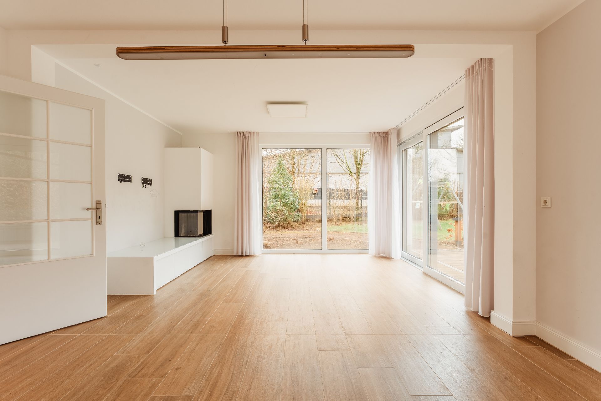 Helles, leeres Wohnzimmer mit Holzfußboden und großen Fenstern zum Garten.