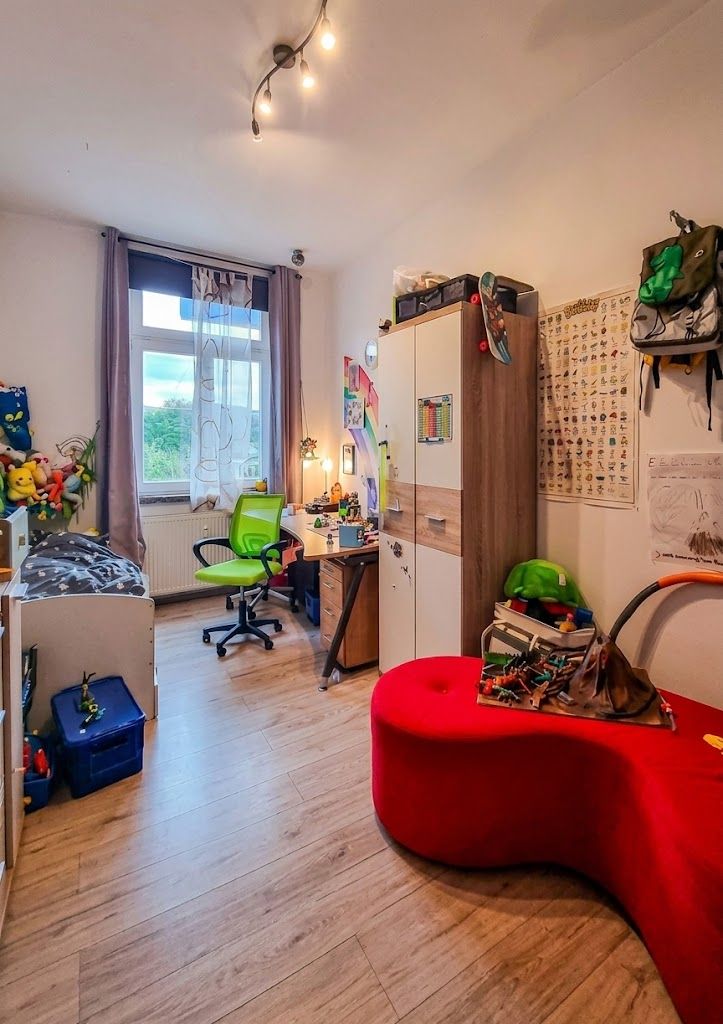 Helles Kinderzimmer mit Bett, Schreibtisch, grünem Stuhl, Schrank und rotem Sofa.