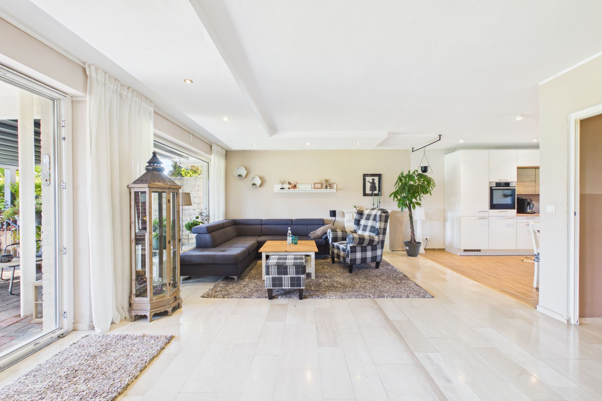 Helles Wohnzimmer mit Ecksofa, Sessel, Couchtisch und offener Küche, große Fenster und Pflanzen.