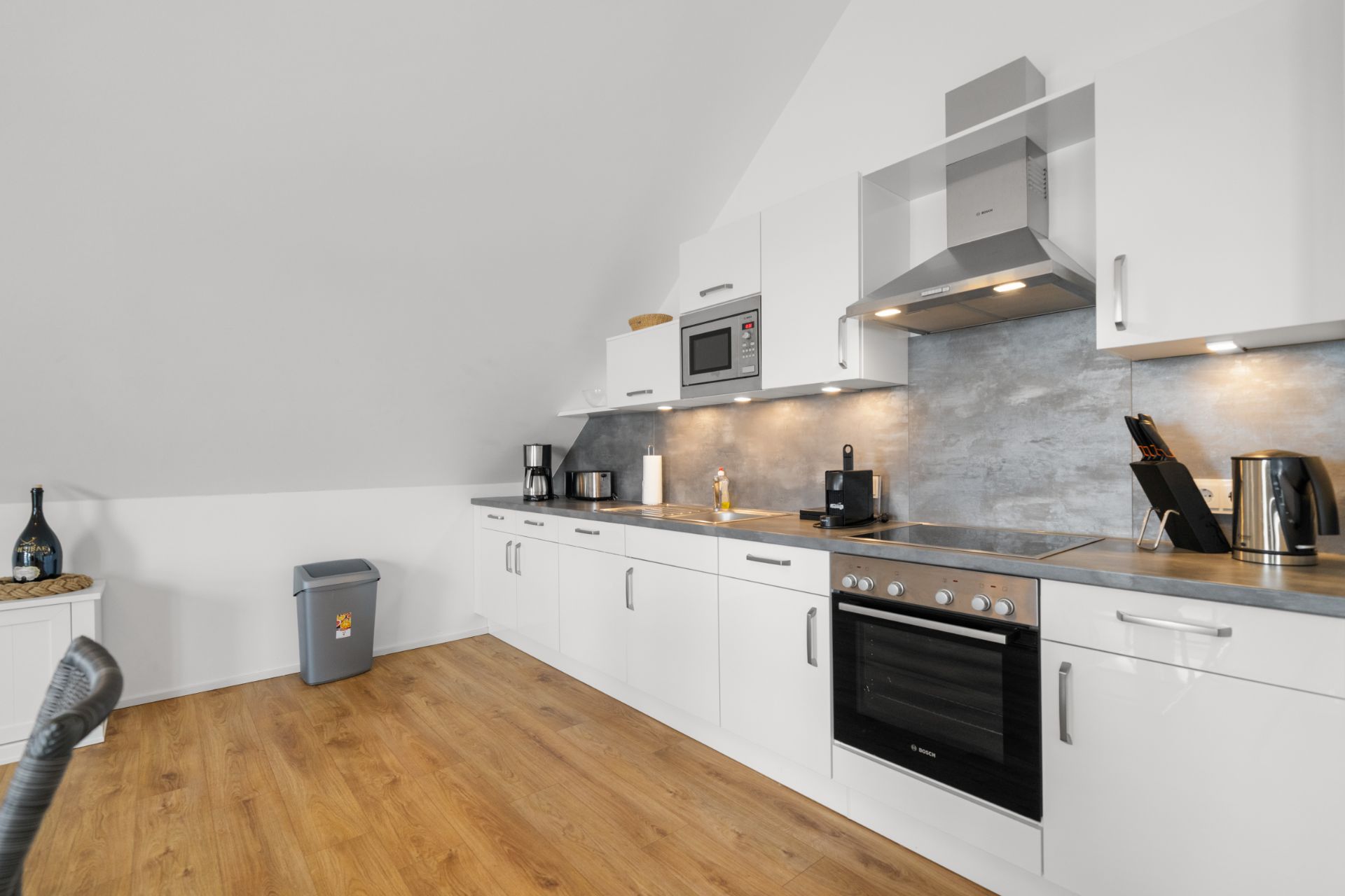 Moderne Küche mit weißen Schränken, Elektrogeräten und Holzfußboden unter Dachschräge.