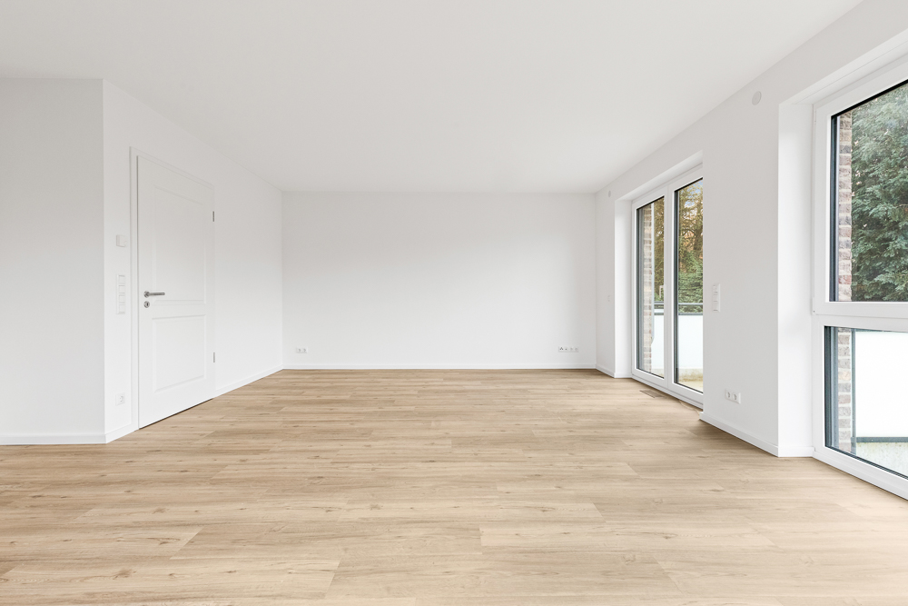 Helles, leeres Zimmer mit Holzboden, weißen Wänden und großen Fenstern.