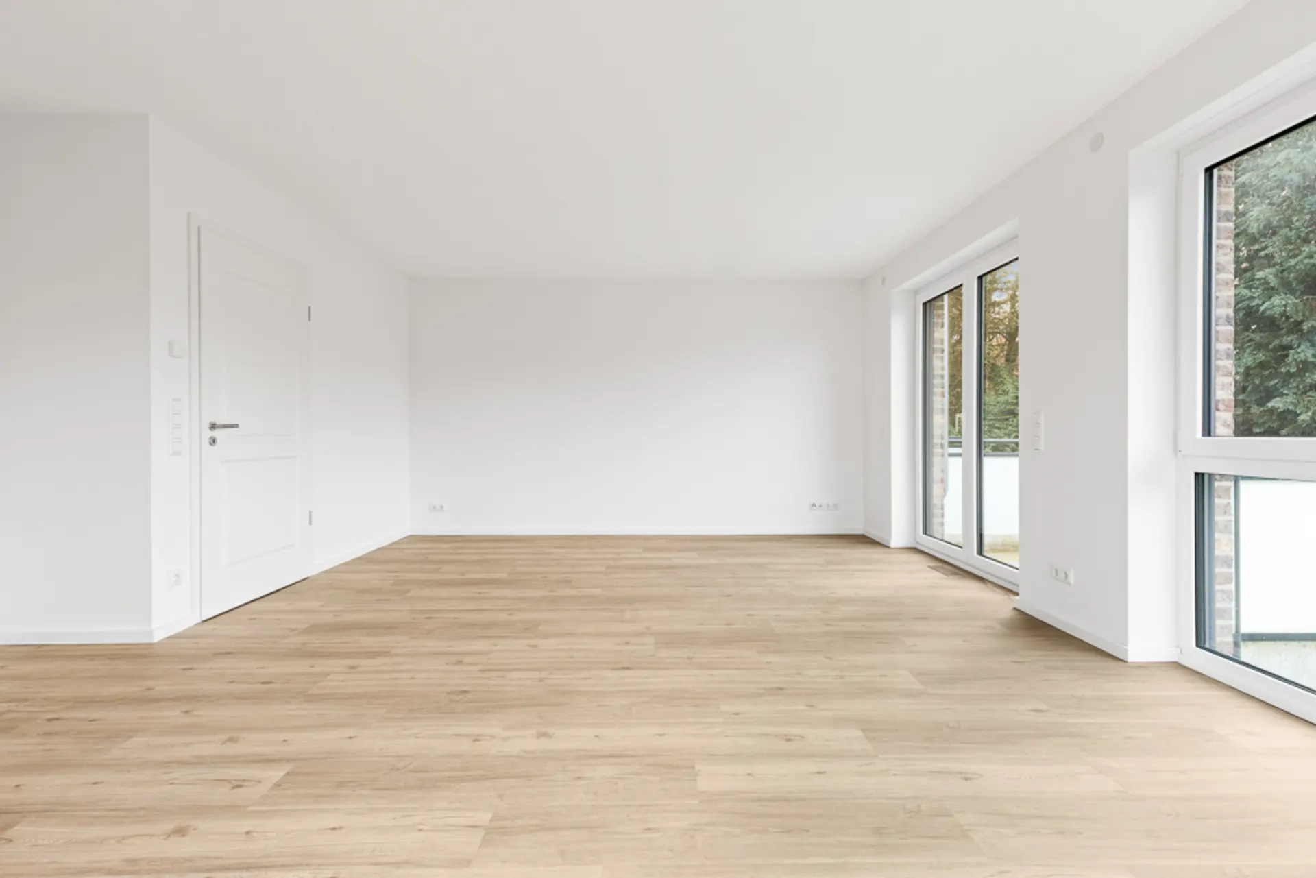 Helles, leeres Zimmer mit Holzboden, weißen Wänden und großen Fenstern.