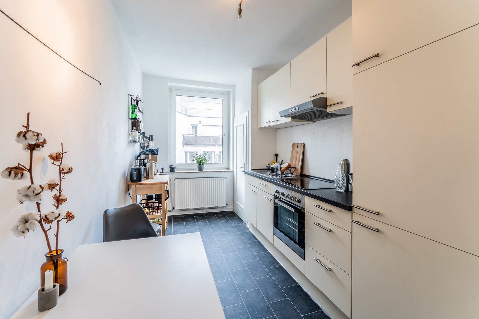 Moderne, helle Küche mit weißen Schränken, Esstisch, Fenster und schwarzen Bodenfliesen.
