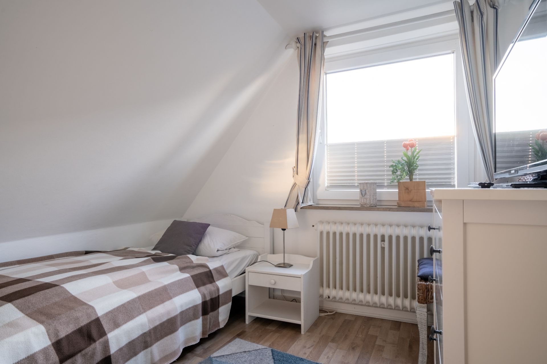 Kleines Schlafzimmer mit Einzelbett, Nachttisch, Fenster und Vorhängen.
