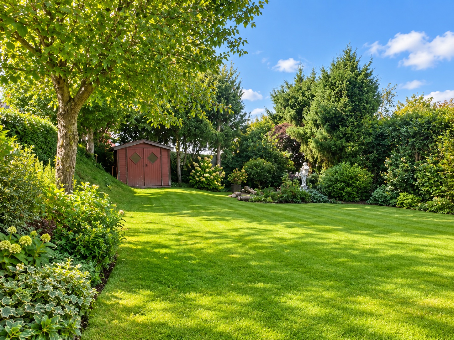 Gepflegter Garten mit grünem Rasen, Bäumen, Sträuchern und einem roten Geräteschuppen im Hintergrund.