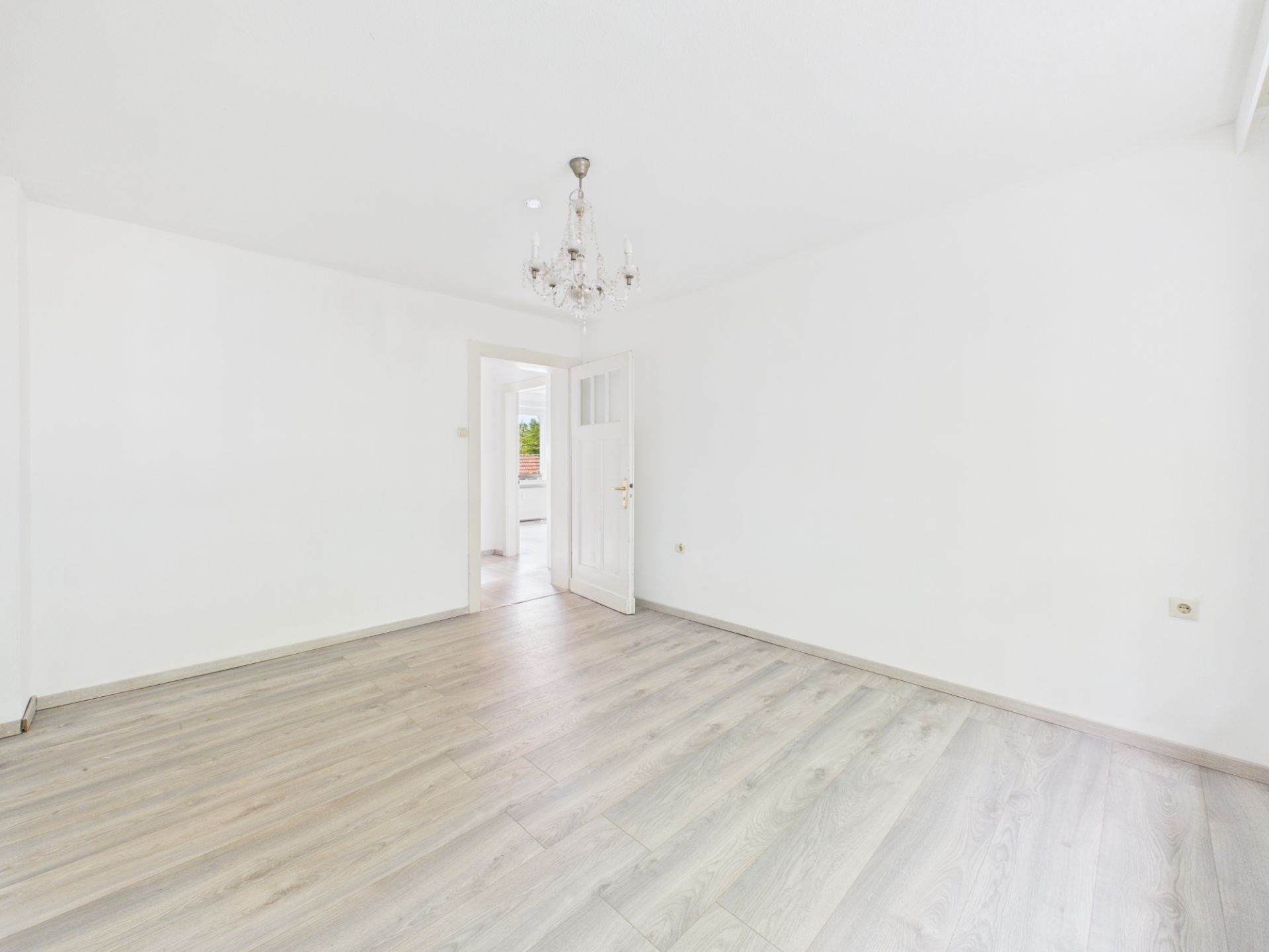 Heller, leerer Raum mit Holzfußboden, weißer Wand und Kronleuchter, offene Tür im Hintergrund.