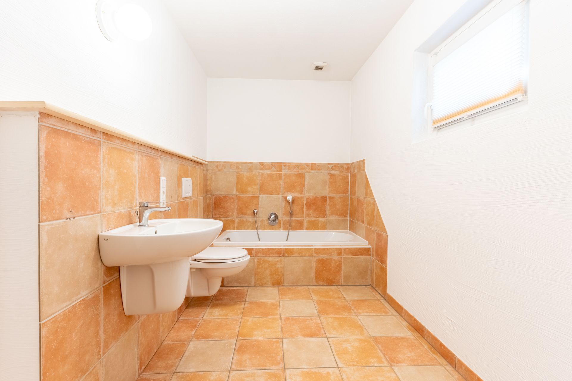 Helles Badezimmer mit Badewanne, Waschbecken und Toilette, orange-gelbe Fliesen, kleines Fenster.