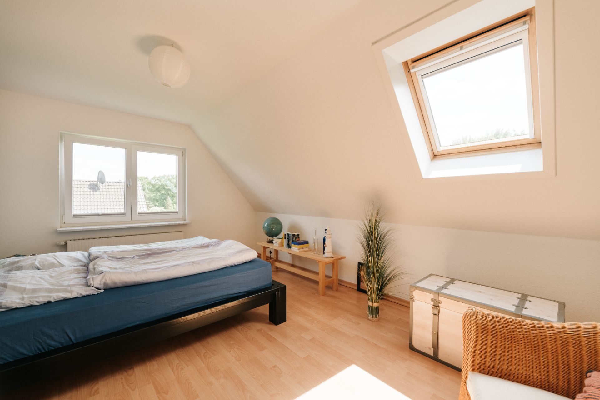 Helles Schlafzimmer mit Bett, Dachfenster, Holzboden, Tisch und Truhe.