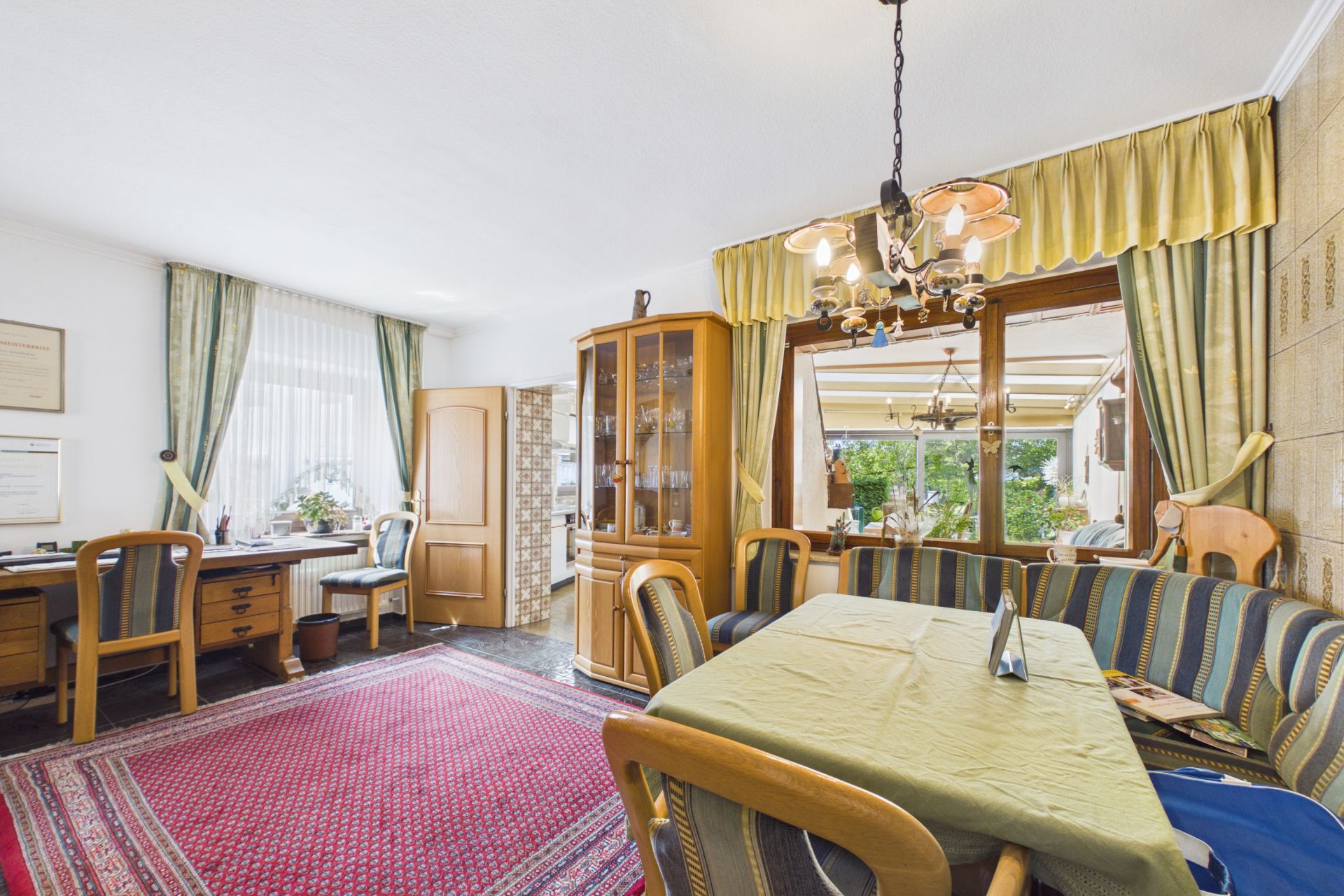 Wohnzimmer mit Esstisch, Eckbank, Schreibtisch, Teppich und großem Fenster mit Vorhängen.