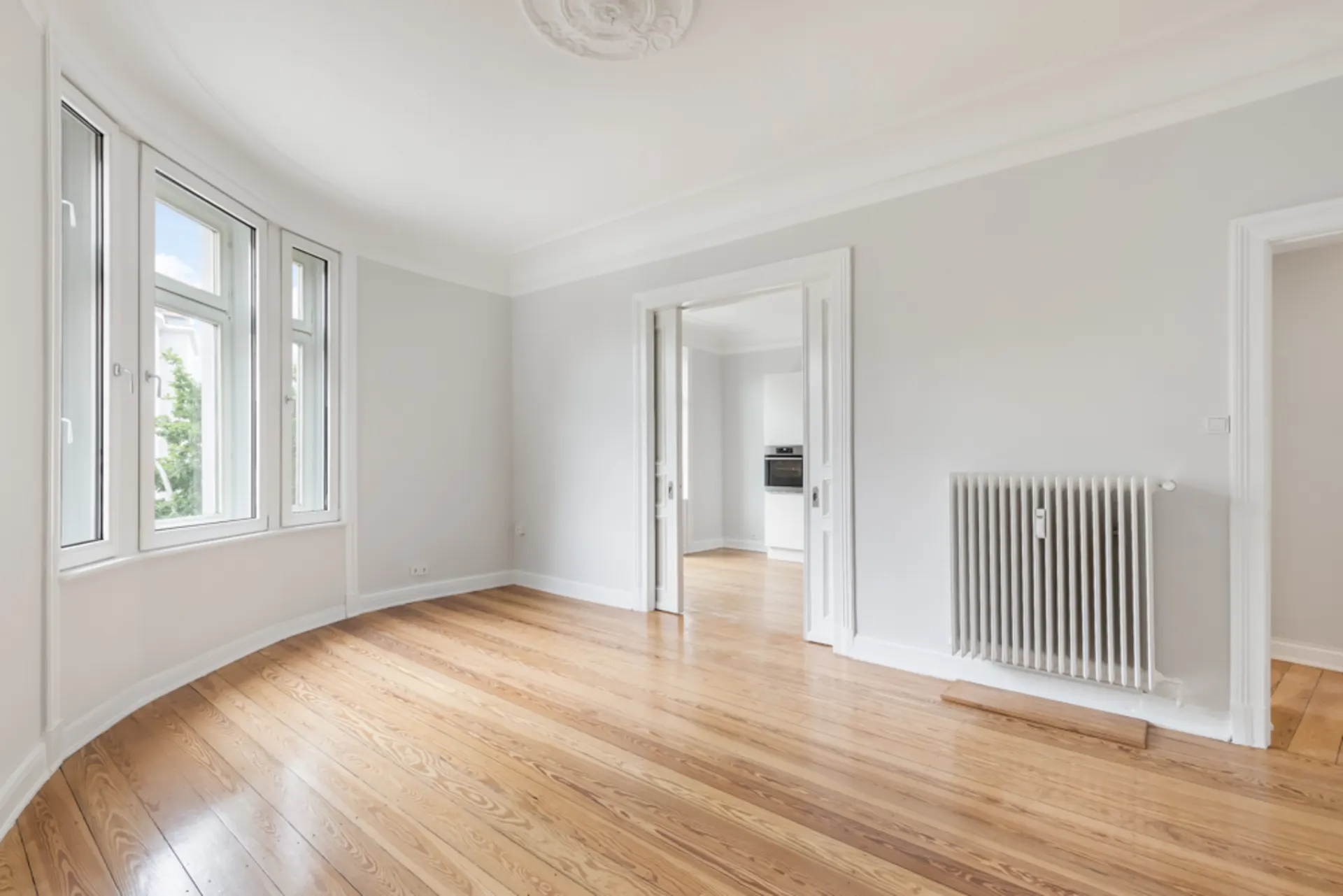 Helles Wohnzimmer mit Holzboden, weißen Wänden und großen Fenstern. Heizkörper an der Wand.