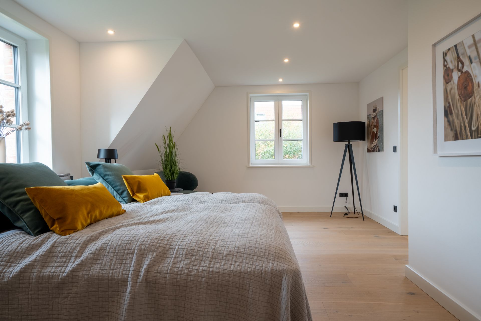 Helles Schlafzimmer mit großem Bett, bunten Kissen, Fenster, Pflanzen und Stehlampe.