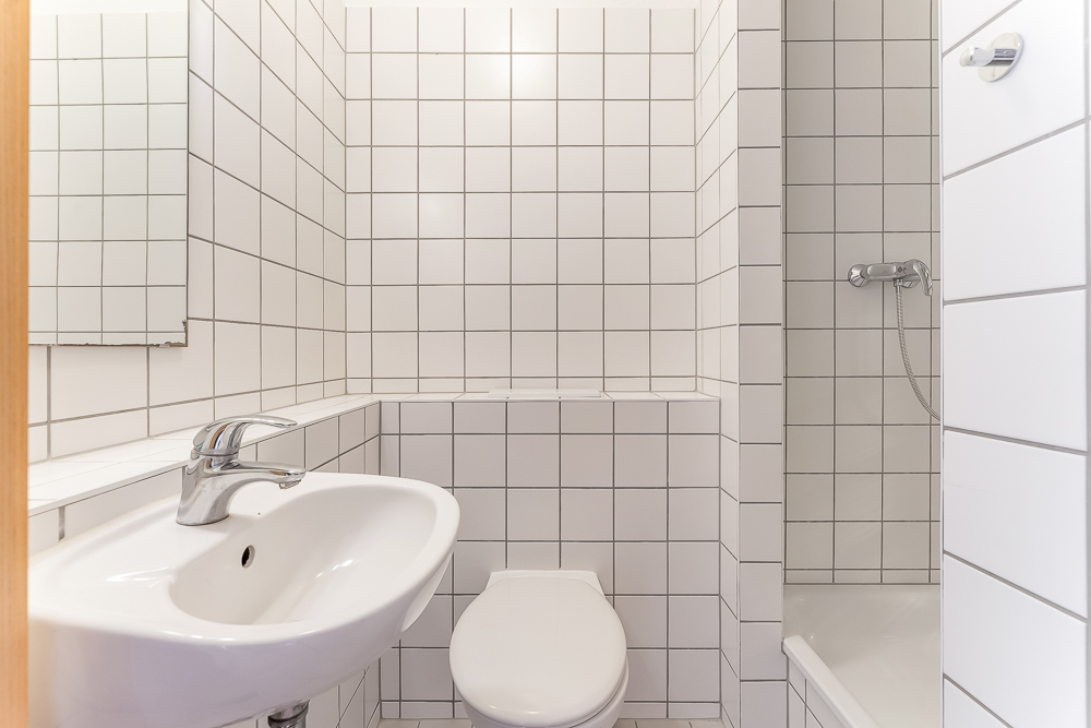 Kleines Bad mit weiß gefliesten Wänden, Waschbecken, Toilette und Dusche.