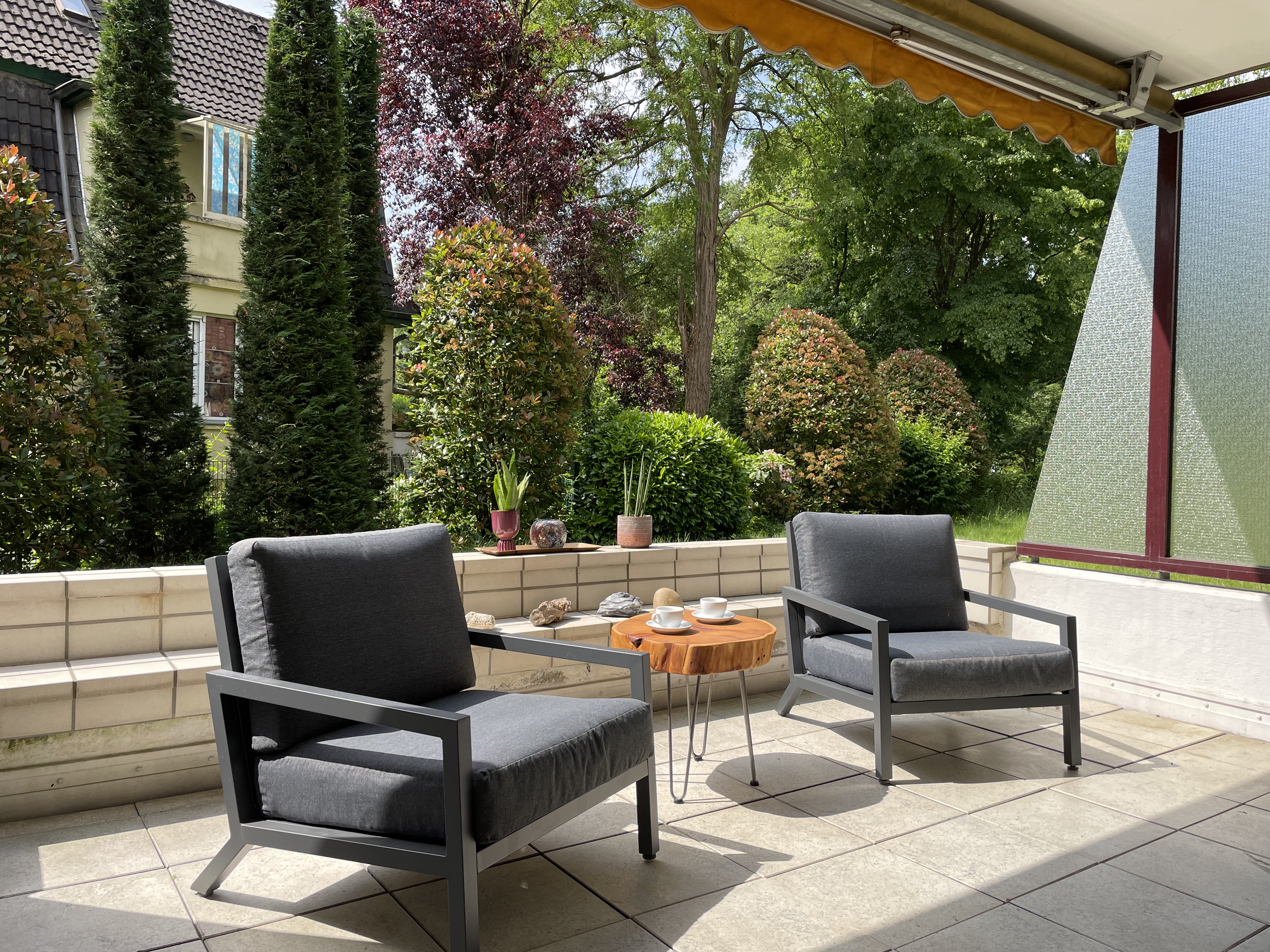 Terrasse mit zwei grauen Sesseln, kleinem Tisch und Blick auf Garten mit Bäumen.