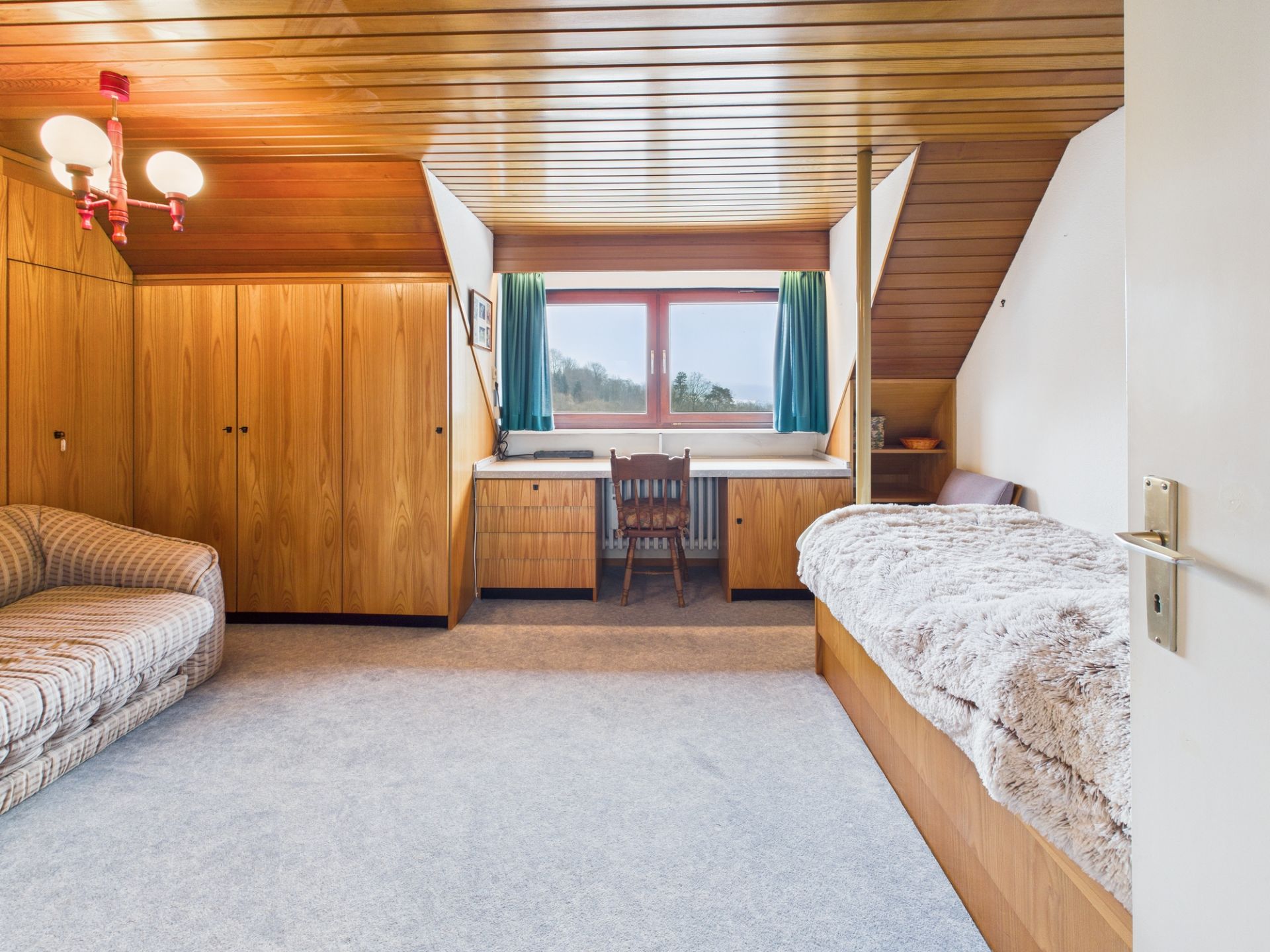Zimmer mit Holzwänden, Sofa, Bett, Schreibtisch am Fenster und Einbauschränken.