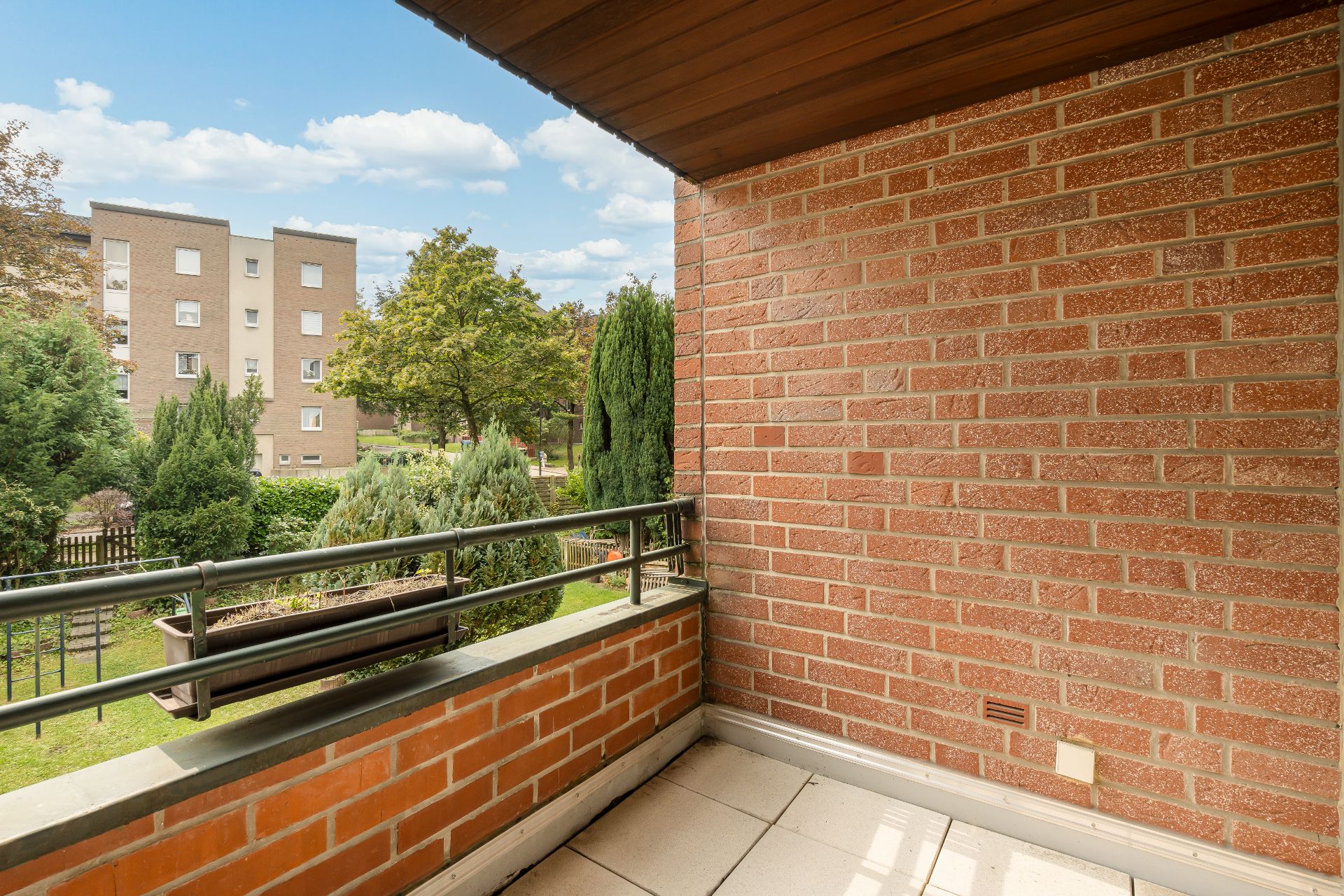Kleiner Balkon mit roter Backsteinwand und Blick auf Grünfläche mit Bäumen.