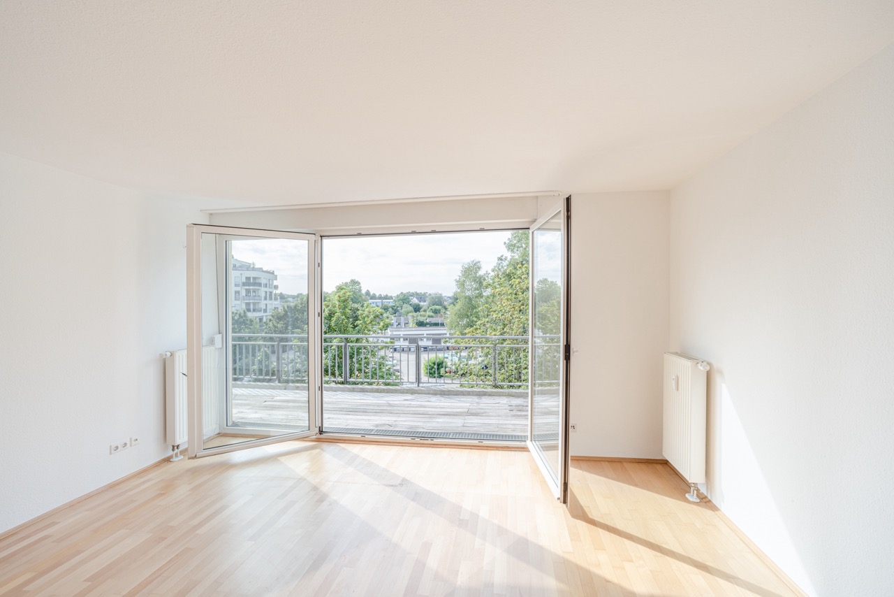 Helles Zimmer mit Parkettboden und großen Fenstern, die zu einem Balkon mit Blick ins Grüne führen.