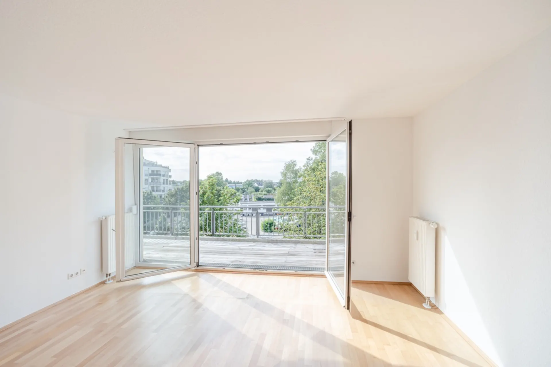 Helles Zimmer mit Parkettboden und großen Fenstern, die zu einem Balkon mit Blick ins Grüne führen.