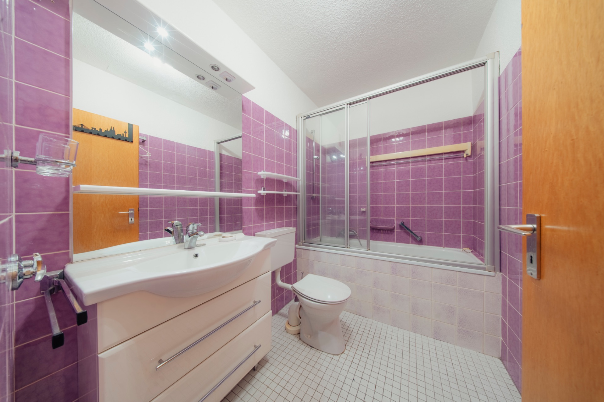 Badezimmer mit lila Fliesen, Waschbecken, Toilette und Badewanne mit Glastrennwand.