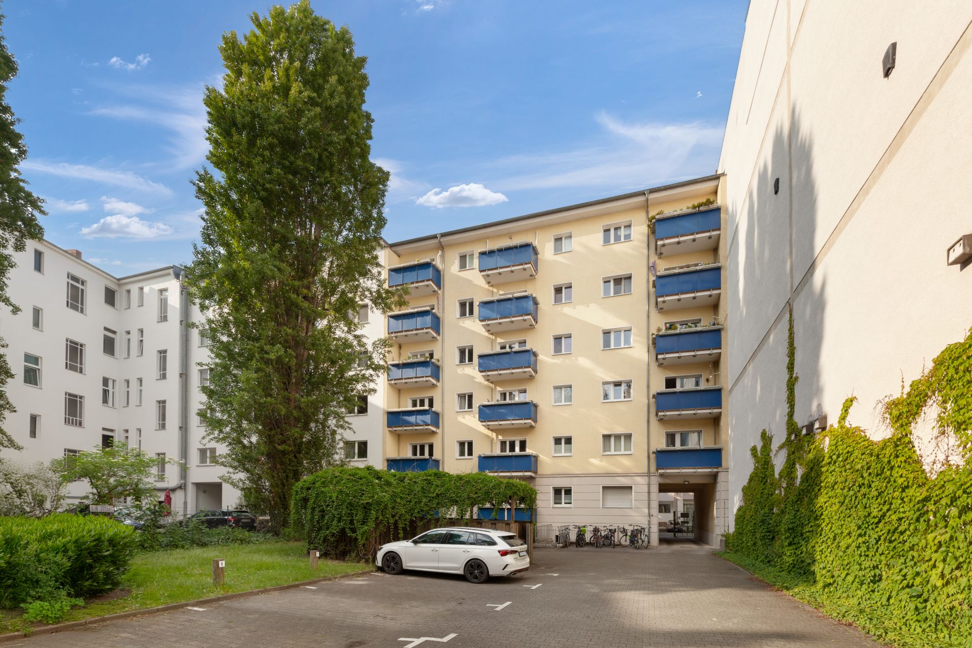 Wohnhaus mit mehreren Balkonen, ein geparkter weißer Wagen und Fahrräder im Hof.
