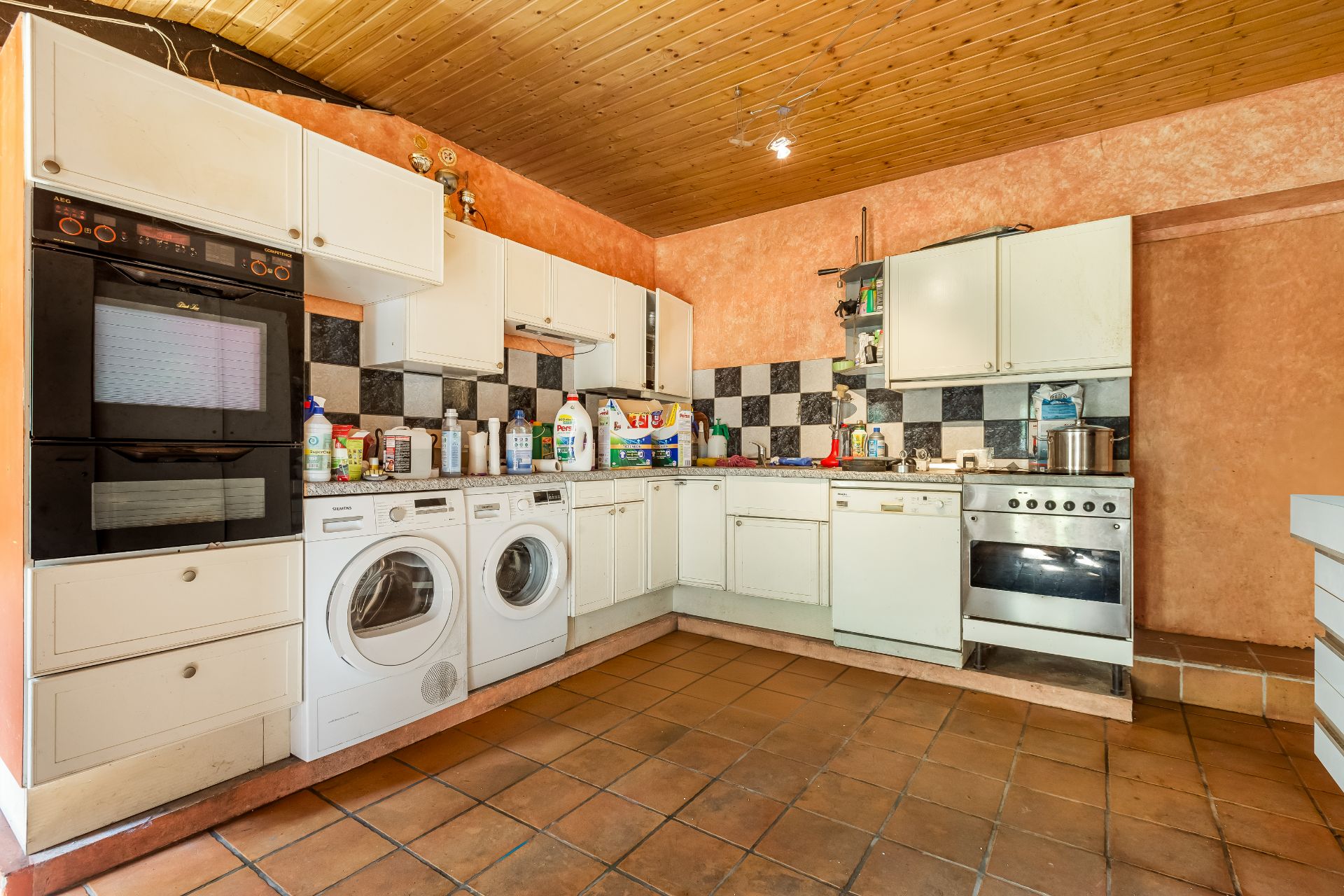 Küche mit weißen Schränken, zwei Waschmaschinen, Herd und Backofen, Fliesenboden und Holzdecke.