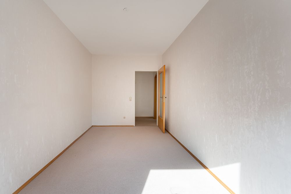 Leeres, schmales Zimmer mit weiß gestrichenen Wänden und offen stehender Holztür.