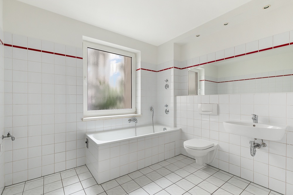 Helles Badezimmer mit Badewanne, Fenster, Wandspiegel, WC und Waschbecken, weiße Fliesen mit rotem Streifen.