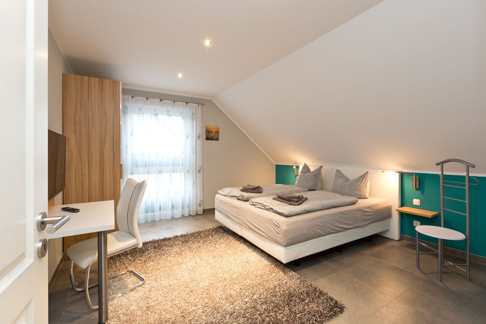 Modernes Schlafzimmer mit Doppelbett, Schreibtisch, Stuhl und großem Fenster mit Vorhang.