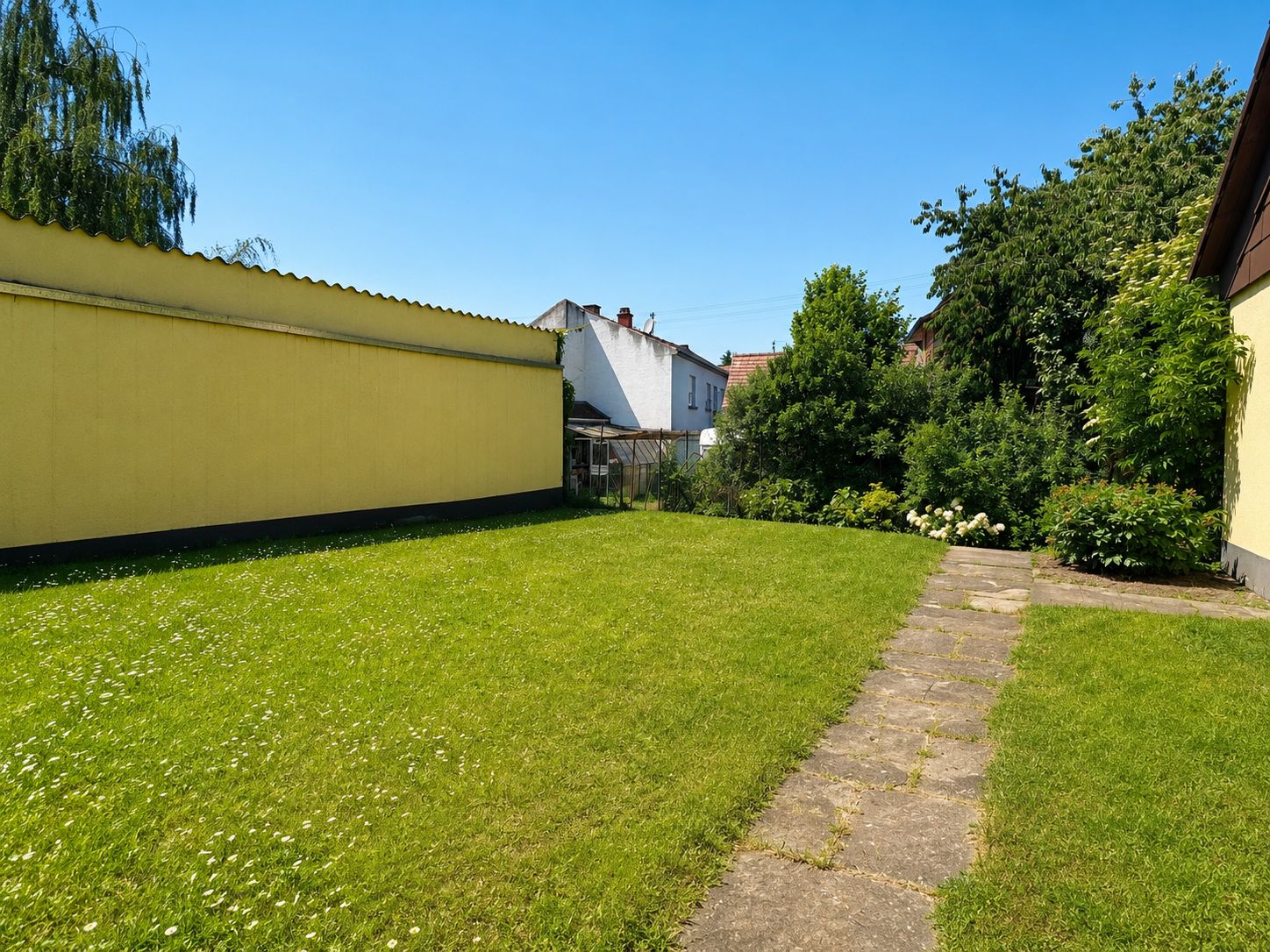 Grüner Garten mit Rasen, Steinweg und gelber Wand, umgeben von Büschen und Bäumen.