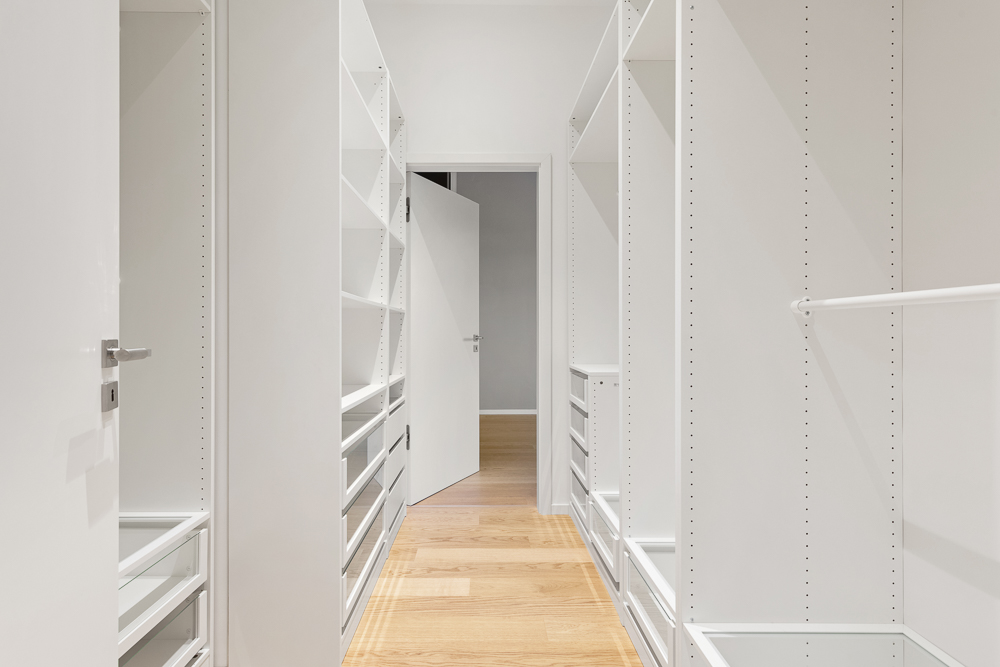 Begehbarer Kleiderschrank mit weißen Regalen und Schubladen, hellem Holzboden und offener Tür.