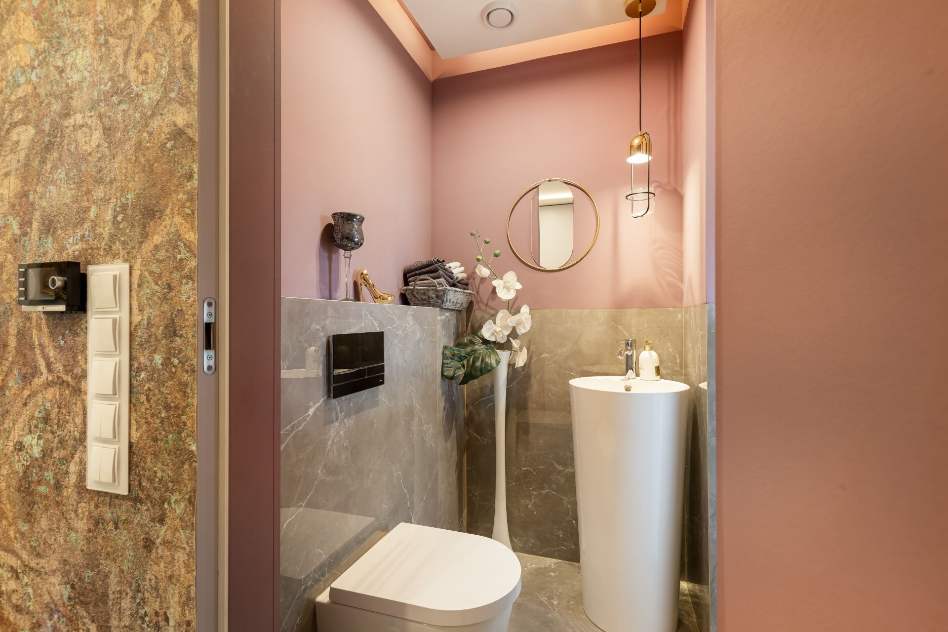 Kleines Badezimmer mit rosafarbenen Wänden, WC, rundem Waschbecken und Spiegel.