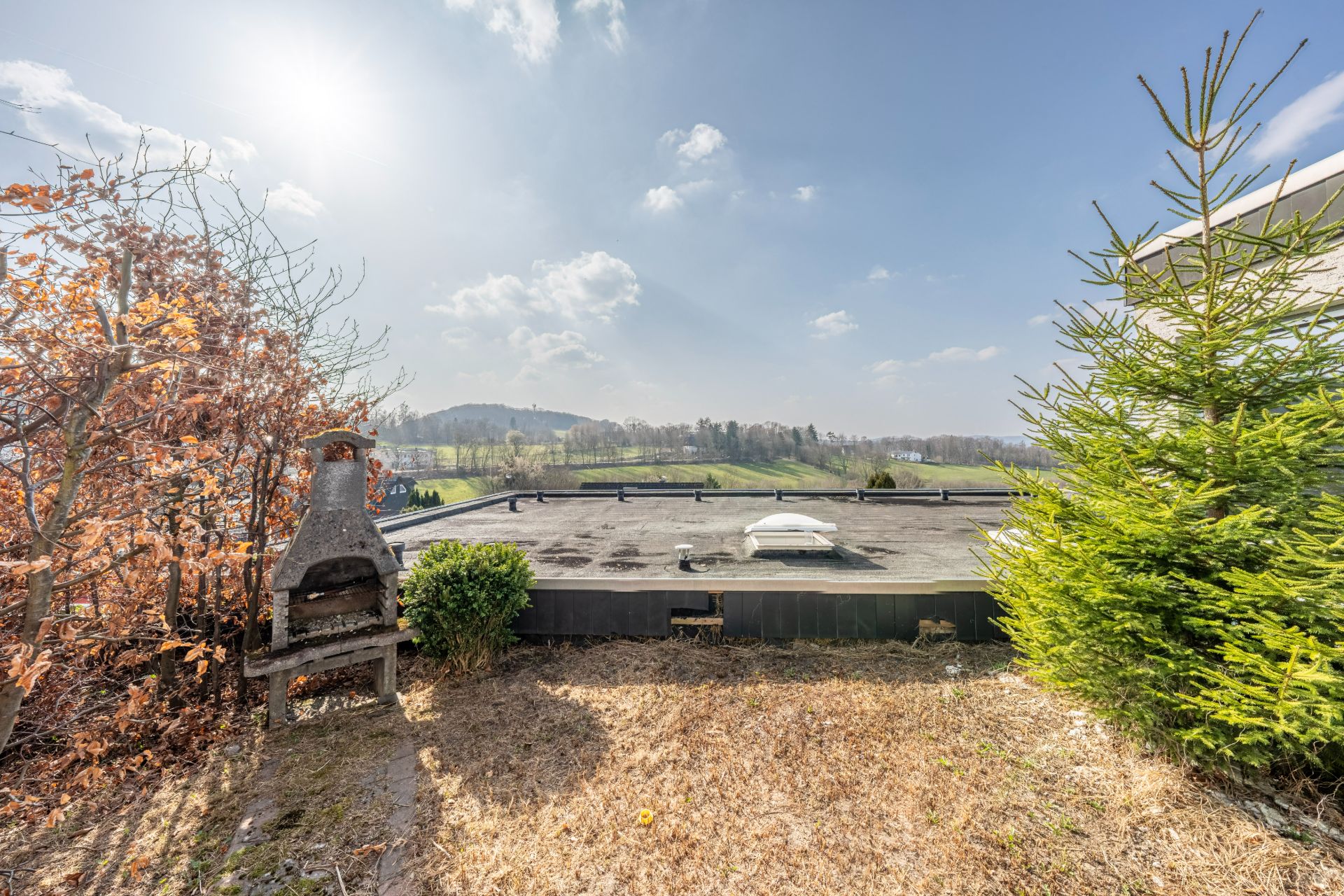 Garten mit Grill, Sträuchern und Blick auf flaches Hausdach sowie Landschaft im Hintergrund.