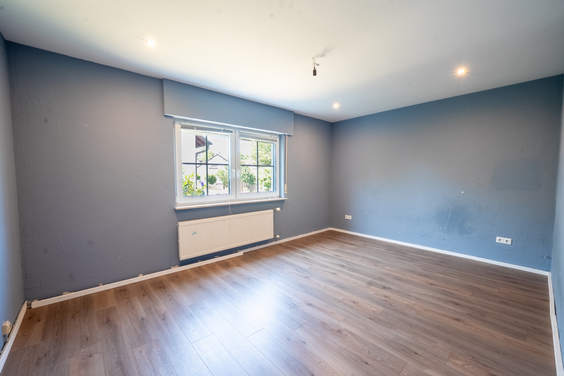 Leerer Raum mit hellblau gestrichenen Wänden, großem Fenster und Holzfußboden.