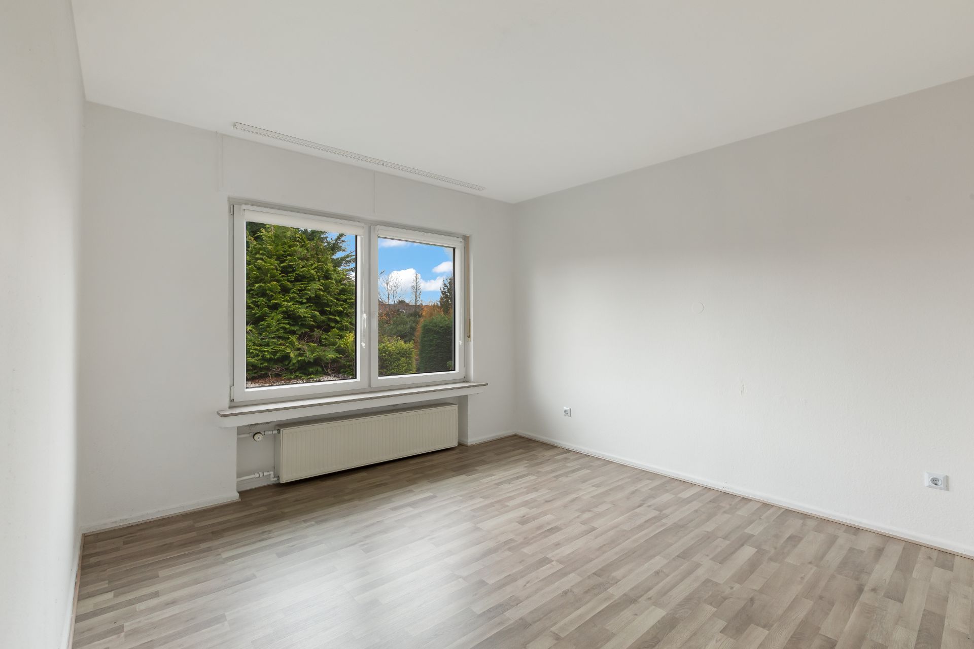 Leeres Zimmer mit hellem Laminatboden und großem Fenster mit Blick ins Grüne.