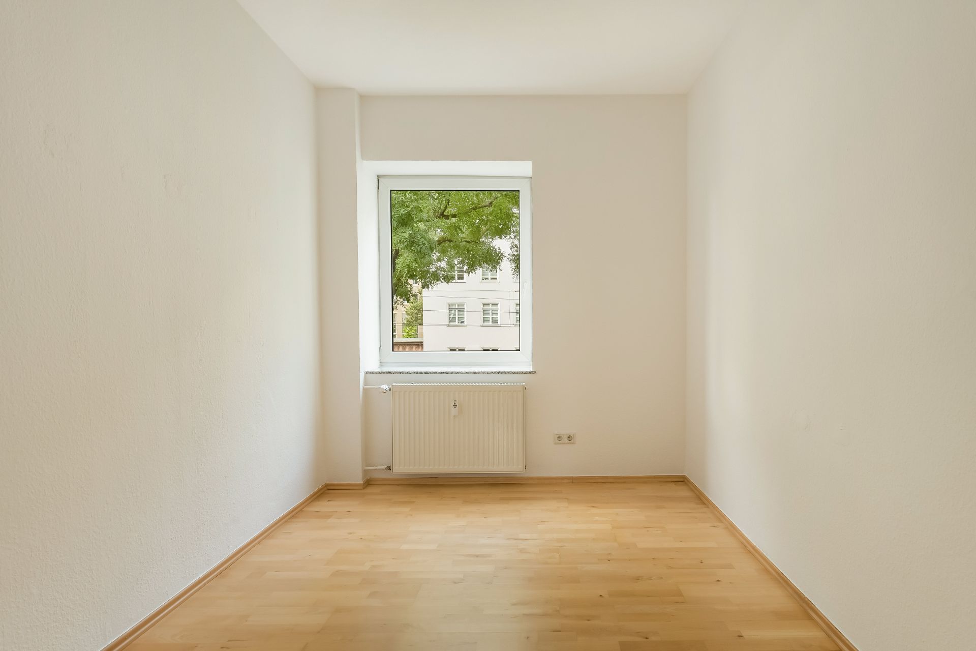 Leerer Raum mit hellem Holzboden, weißer Wand und Fenster mit Blick auf einen Baum.