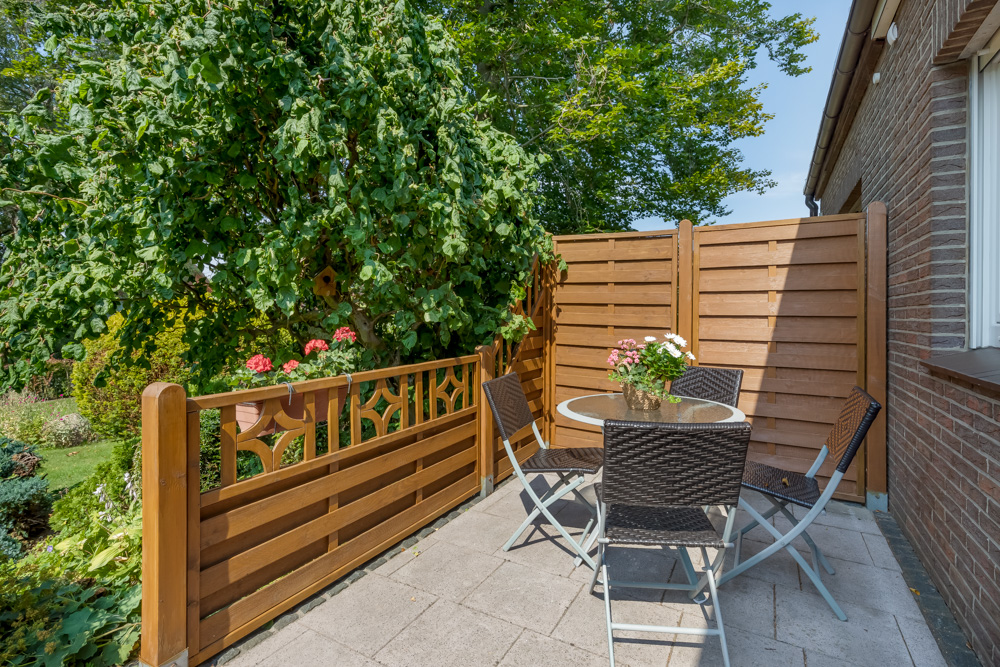 Kleine Terrasse mit Glastisch, vier Stühlen, Holzzaun und Pflanze neben einem Wohnhaus.
