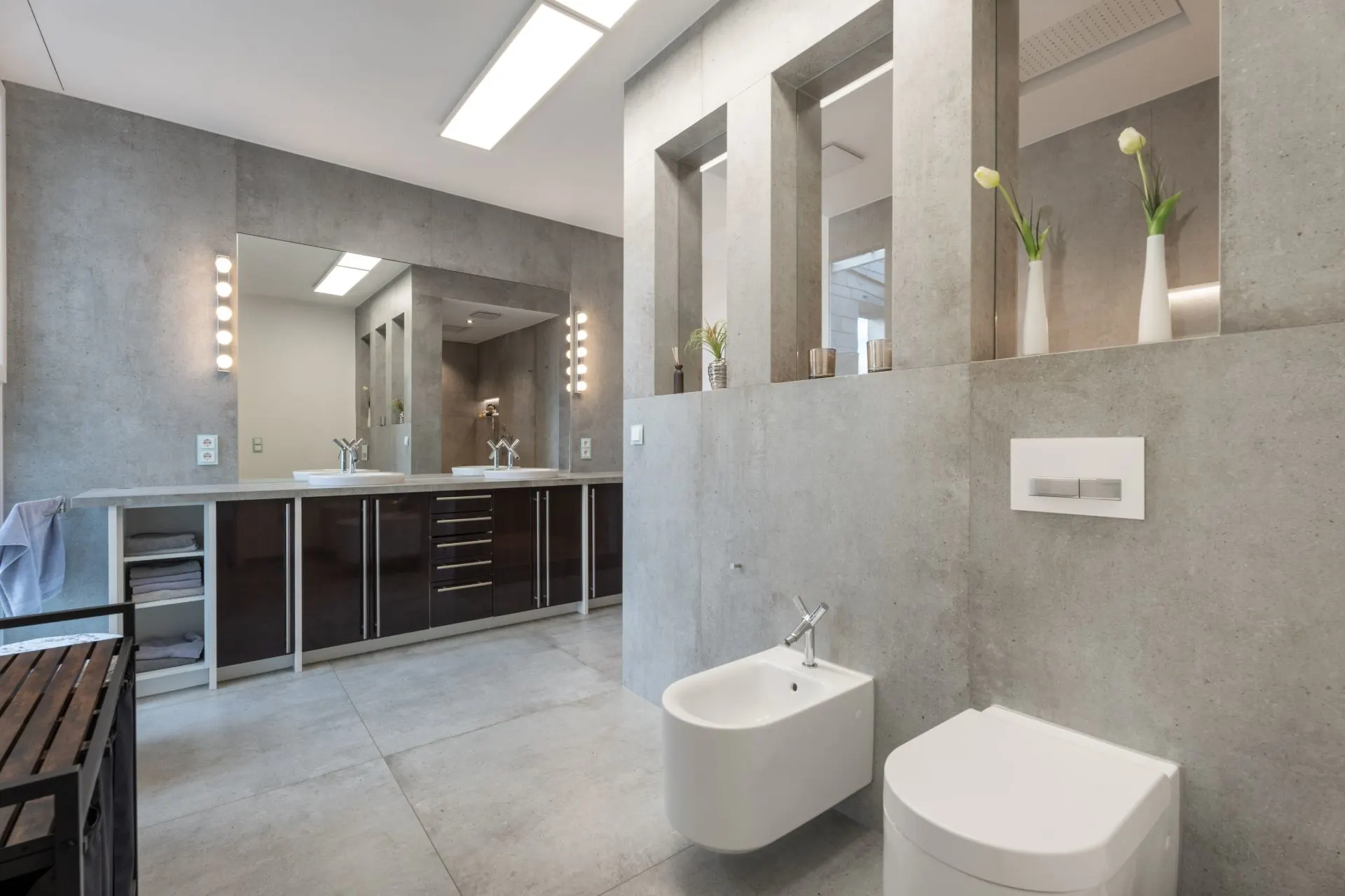 Modernes Badezimmer mit grauen Fliesen, WC, Bidet, großem Spiegel und Dekovasen auf einer Trennwand.