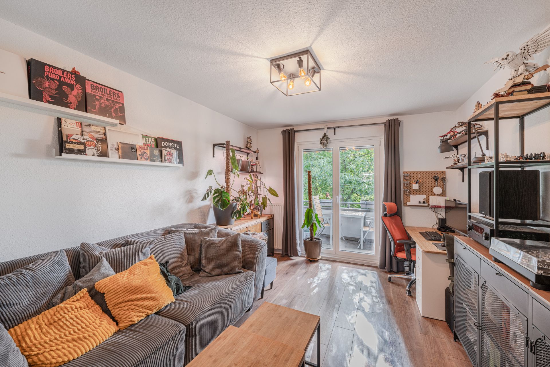 Wohnzimmer mit grauem Sofa, Schreibtisch, Regal, Pflanzen und Zugang zu einem Balkon.